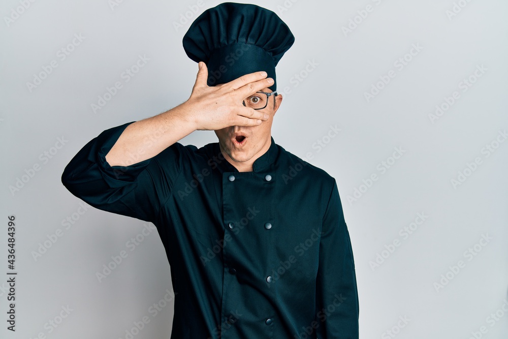 Handsome young man wearing professional cook uniform and hat peeking in shock covering face and eyes with hand, looking through fingers afraid