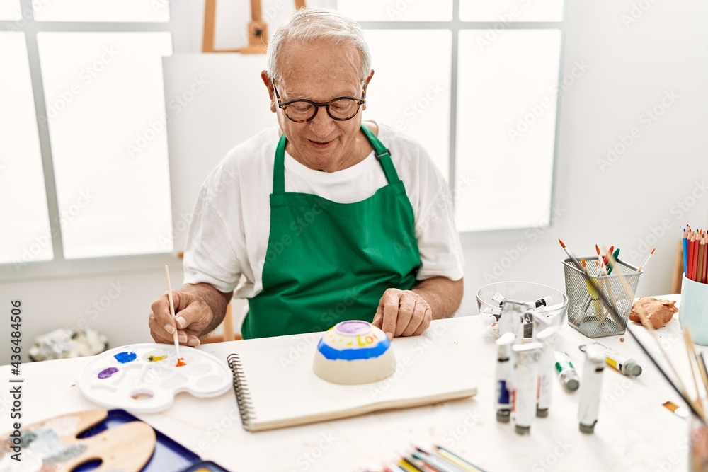 Senior grey-haired artist man smiling happy painting pottery sitting on the table at art studio. Senior grey-haired artist man smiling happy painting pottery sitting on the table at art studio.