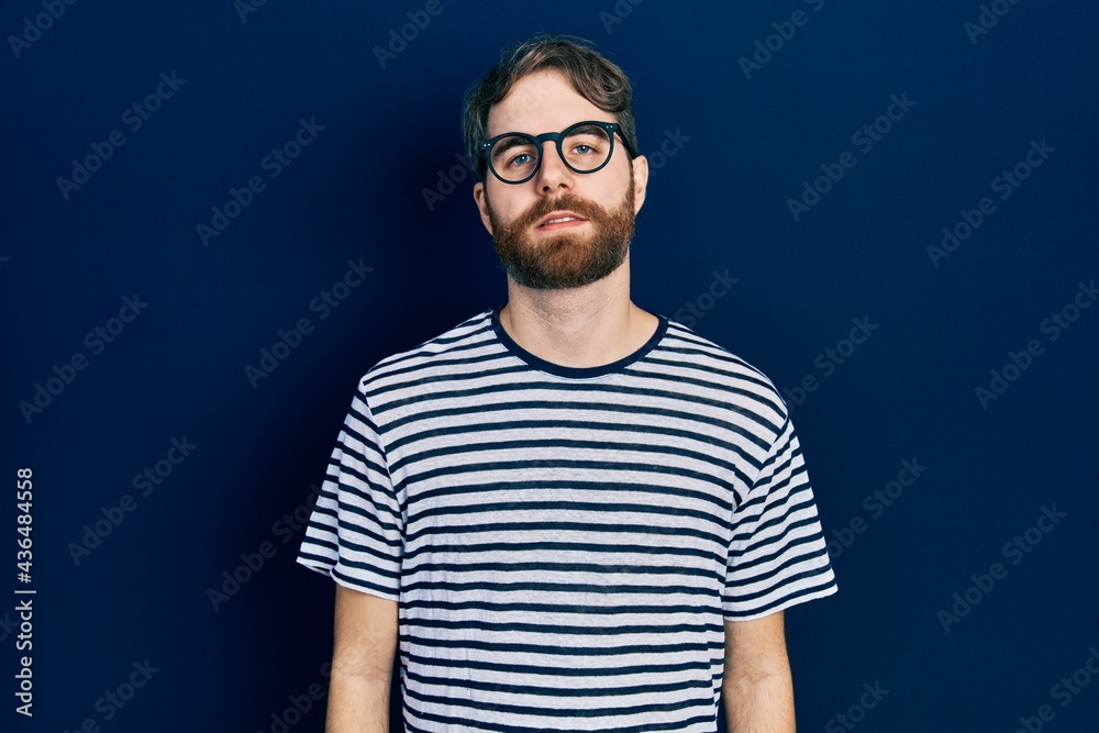 Caucasian man with beard wearing striped t shirt and glasses relaxed with serious expression on face. simple and natural looking at the camera.