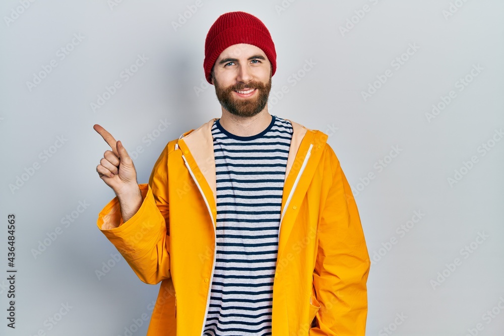 Caucasian man with beard wearing yellow raincoat with a big smile on face, pointing with hand finger to the side looking at the camera. Caucasian man with beard wearing yellow raincoat with a big smile on face, pointing with hand finger to the side looking at the camera.