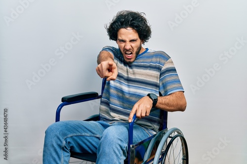 Handsome hispanic man sitting on wheelchair pointing displeased and frustrated to the camera, angry and furious with you