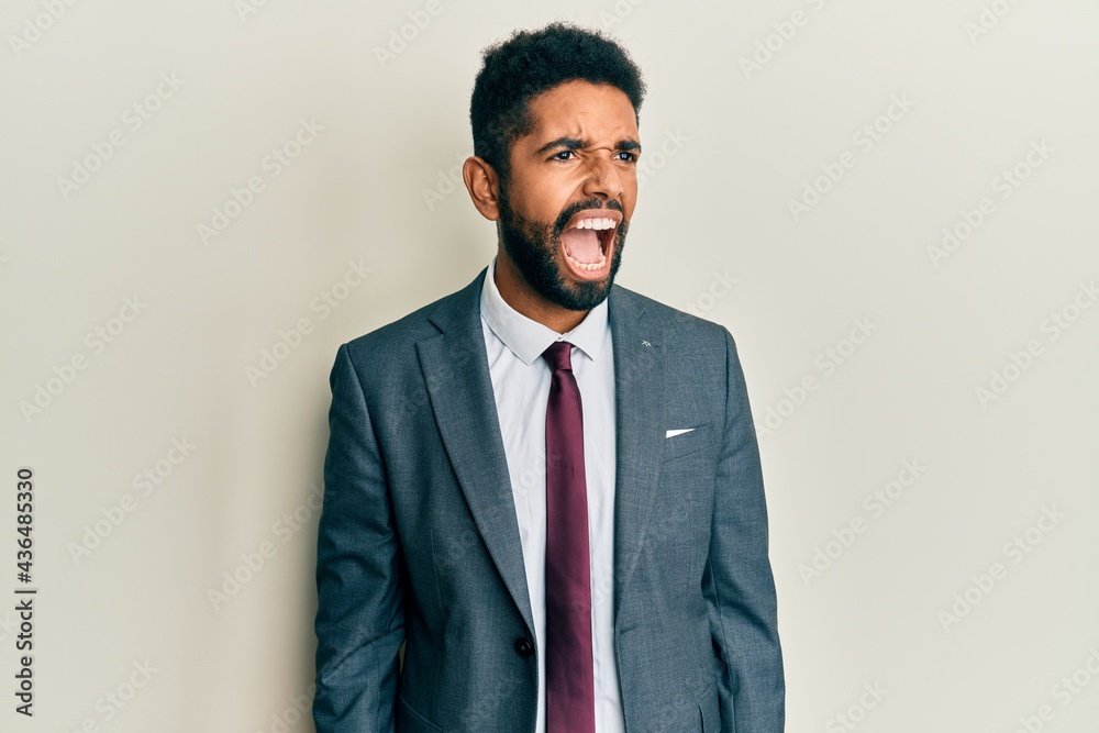 Handsome hispanic man with beard wearing business suit and tie angry and mad screaming frustrated and furious, shouting with anger. rage and aggressive concept.