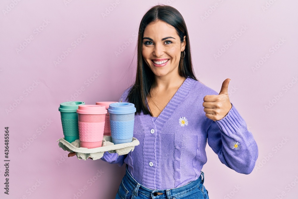 Young brunette woman holding tray with take away coffee smiling happy and positive, thumb up doing excellent and approval sign Young brunette woman holding tray with take away coffee smiling happy and positive, thumb up doing excellent and approval sign