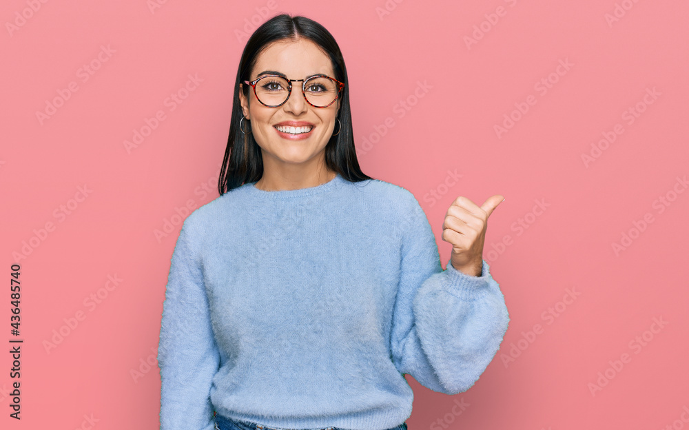 Young hispanic woman wearing casual clothes and glasses smiling with happy face looking and pointing to the side with thumb up. Young hispanic woman wearing casual clothes and glasses smiling with happy face looking and pointing to the side with thumb up.