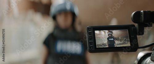 Behind the scenes of female war journalist correspondent wearing bulletproof vest and helmet reporting live near destroyed building, military personnel in the background