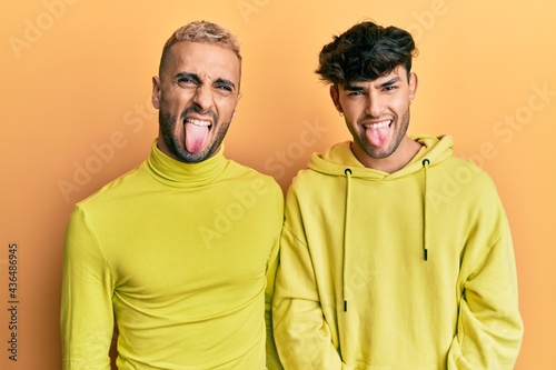 Homosexual gay couple standing together wearing yellow clothes sticking tongue out happy with funny expression. emotion concept.