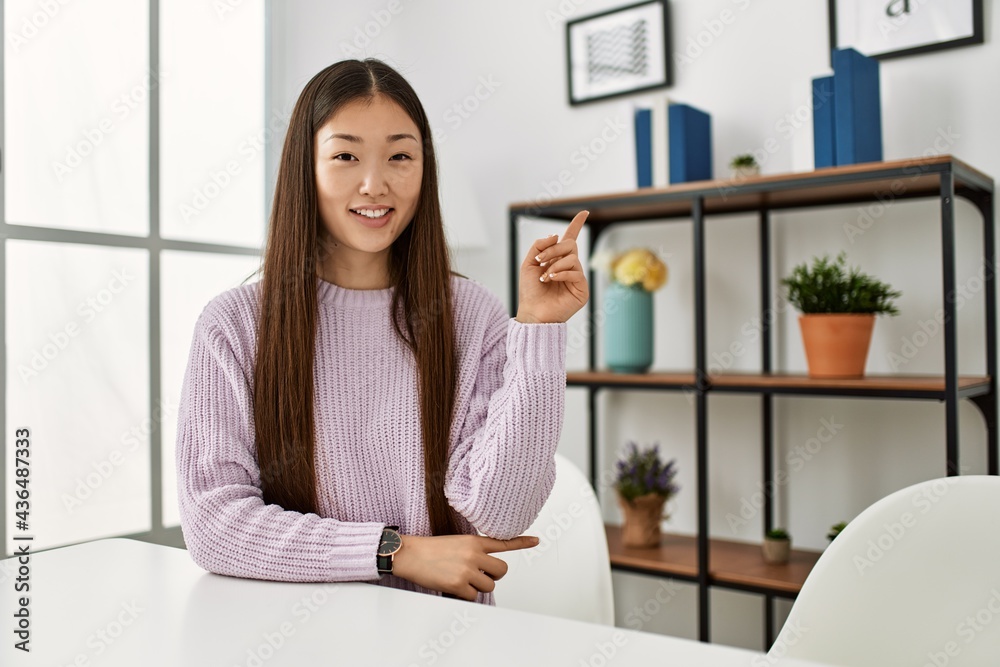 Young chinese girl wearing casual clothes sitting on the table at home smiling happy pointing with hand and finger to the side Young chinese girl wearing casual clothes sitting on the table at home smiling happy pointing with hand and finger to the side