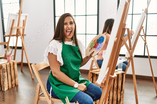 Young hispanic artist women painting on canvas at art studio sticking tongue out happy with funny expression. emotion concept.