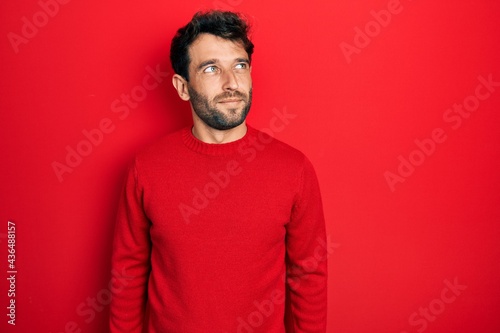 Handsome man with beard wearing casual red sweater smiling looking to the side and staring away thinking.