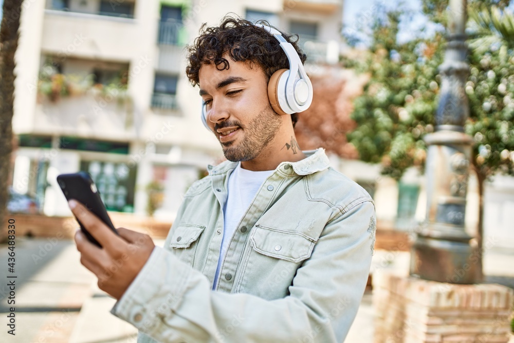 Handsome hispanic man with beard smiling happy outdoors on a sunny day wearing headphones listening to music with smartphone Handsome hispanic man with beard smiling happy outdoors on a sunny day wearing headphones listening to music with smartphone