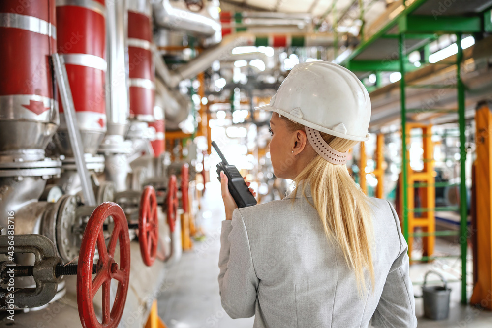 Rear view of blond female supervisor with helmet in suit standing in heating plant and talking over wakie talkie with her employee. Rear view of blond female supervisor with helmet in suit standing in heating plant and talking over wakie talkie with her employee.