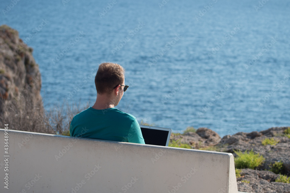 Young man working on a laptop sitting on a bench in a viewpoint in front of the sea. Remote working concept.