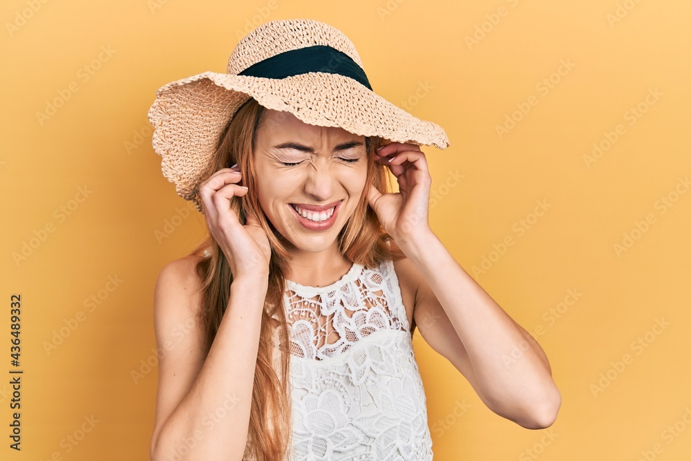 Young caucasian woman wearing summer hat covering ears with fingers with annoyed expression for the noise of loud music. deaf concept.