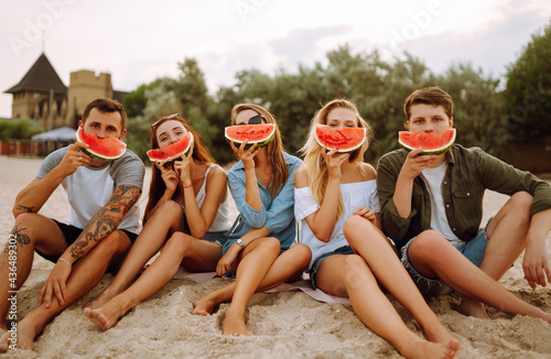 Young friends relaxing on the beach and eating watermelon. Group of people enjoy summer party together. People, lifestyle, travel, nature and vacations concept.