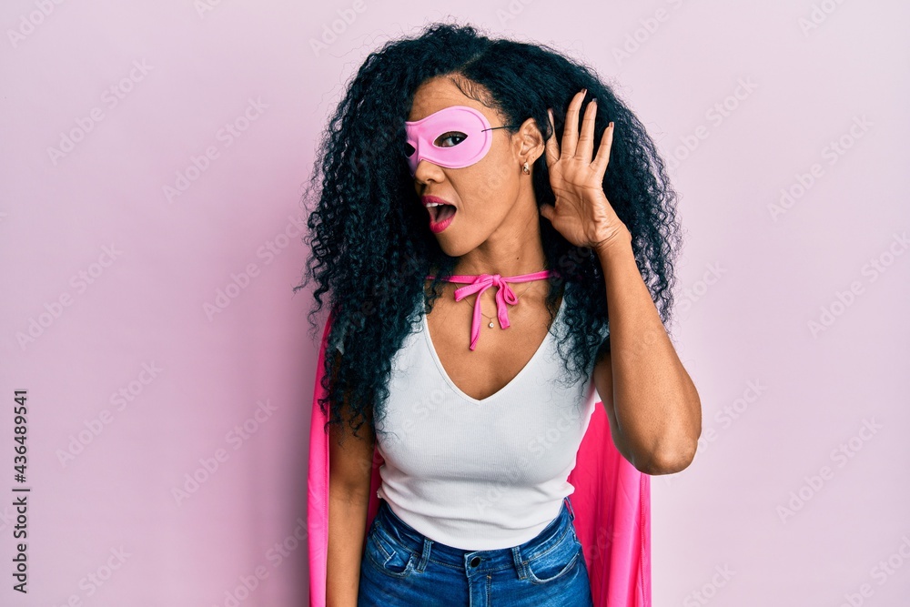 Middle age african american woman wearing super hero costume smiling with hand over ear listening an hearing to rumor or gossip. deafness concept. Middle age african american woman wearing super hero costume smiling with hand over ear listening an hearing to rumor or gossip. deafness concept.
