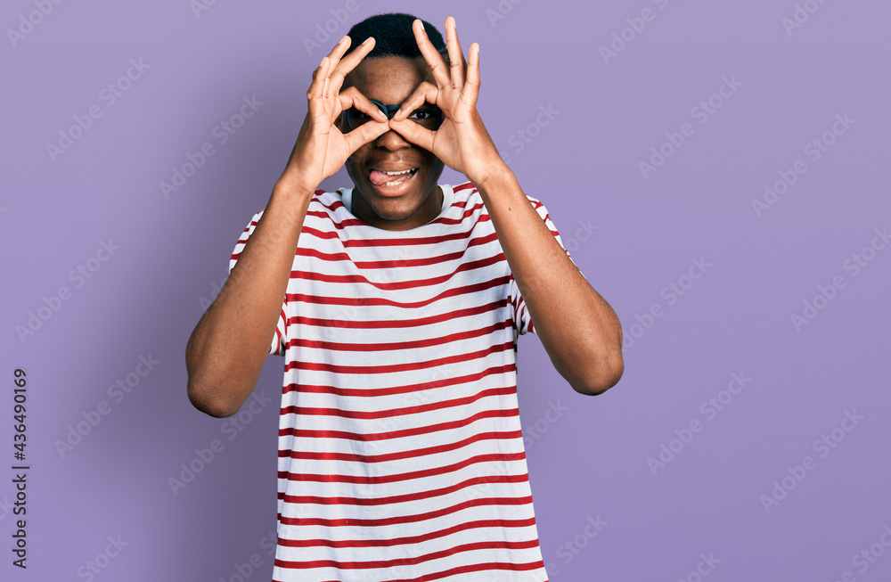 Young african american man wearing casual t shirt and glasses doing ok gesture like binoculars sticking tongue out, eyes looking through fingers. crazy expression. Young african american man wearing casual t shirt and glasses doing ok gesture like binoculars sticking tongue out, eyes looking through fingers. crazy expression.