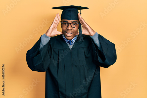 Young african american man wearing graduation cap and ceremony robe smiling cheerful playing peek a boo with hands showing face. surprised and exited