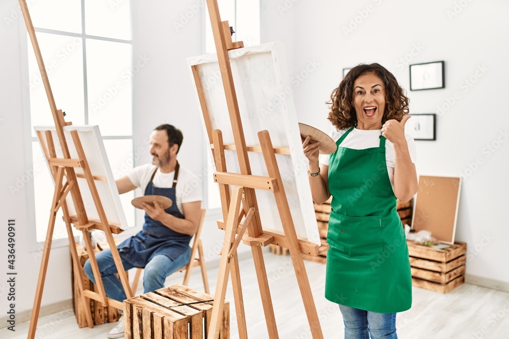 Hispanic middle age woman and mature man at art studio pointing thumb up to the side smiling happy with open mouth Hispanic middle age woman and mature man at art studio pointing thumb up to the side smiling happy with open mouth