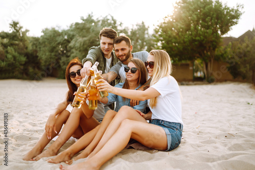 Group of friends cheers and drink beers on the beach. Young friends relaxing on the beath having picnic, toasting with beerus. People, lifestyle, travel, nature and vacations concept.