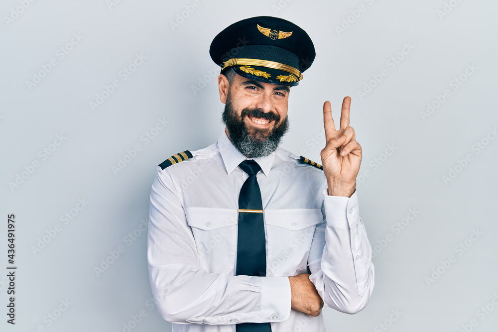 Young hispanic man wearing airplane pilot uniform smiling with happy face winking at the camera doing victory sign. number two. Young hispanic man wearing airplane pilot uniform smiling with happy face winking at the camera doing victory sign. number two.