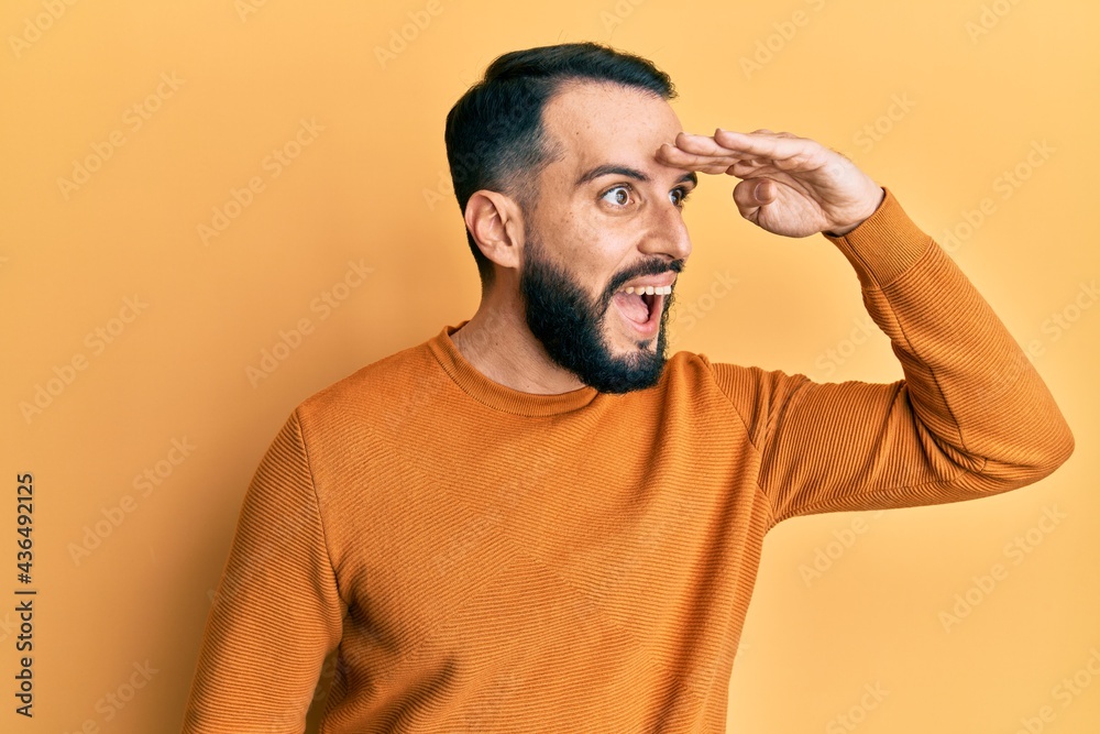 Young man with beard wearing casual winter sweater very happy and smiling looking far away with hand over head. searching concept. Young man with beard wearing casual winter sweater very happy and smiling looking far away with hand over head. searching concept.