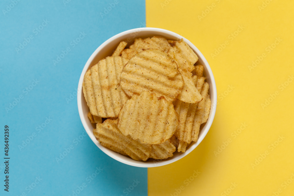 Close up of Wrinkled Wavy Potato Chips in white ceramic bowl, Popular Ready to eat crunchy, salty pale-yellow color over blue yellow background Close up of Wrinkled Wavy Potato Chips in white ceramic bowl, Popular Ready to eat crunchy, salty pale-yellow color over blue yellow background