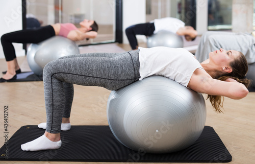 Young woman maintaining active lifestyle exercising with gym ball during group pilates class in modern fitness center