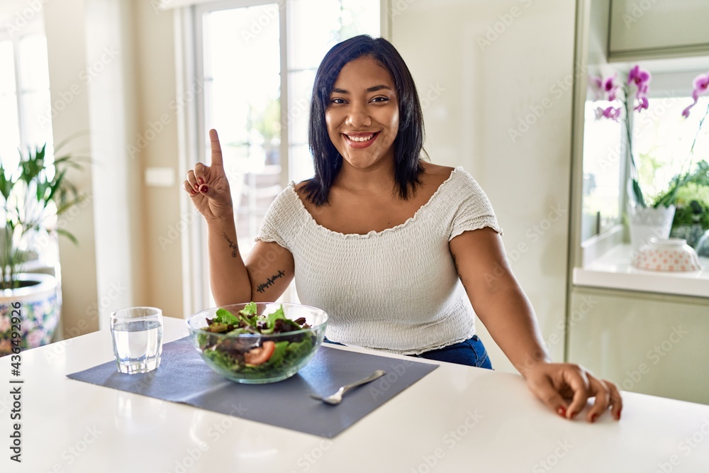Young hispanic woman eating healthy salad at home showing and pointing up with finger number one while smiling confident and happy.