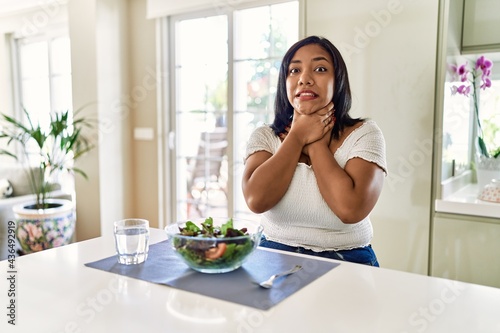 Young hispanic woman eating healthy salad at home shouting and suffocate because painful strangle. health problem. asphyxiate and suicide concept.