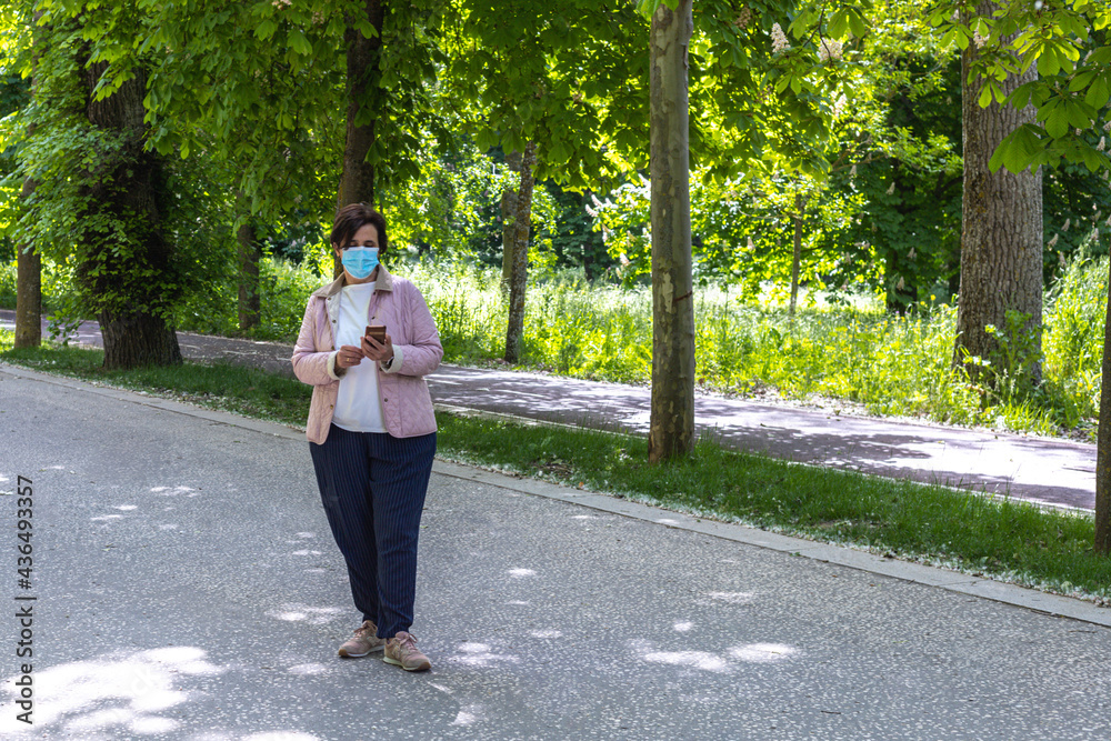 Woman with mask, jacket and pink slippers using smart phone in the middle of the road with trees in the background and copy space