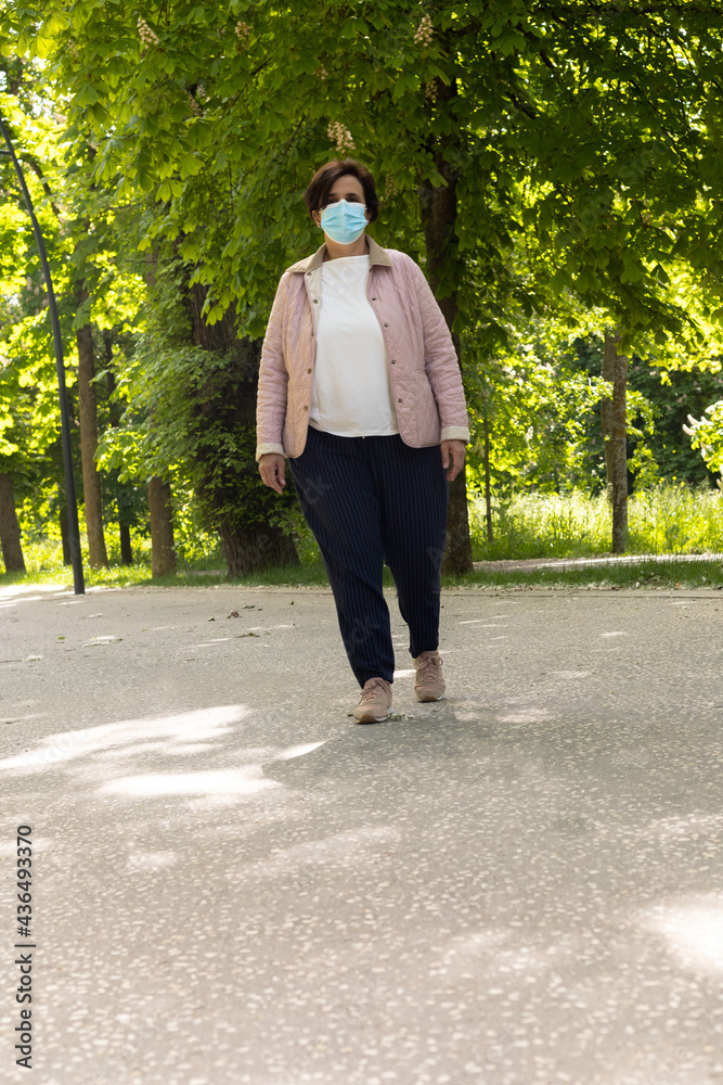 Fototapeta premium Vertical photo with copy space of a middle-aged woman walking with a mask, pink jacket and pink slippers on a sunny day with trees in the background
