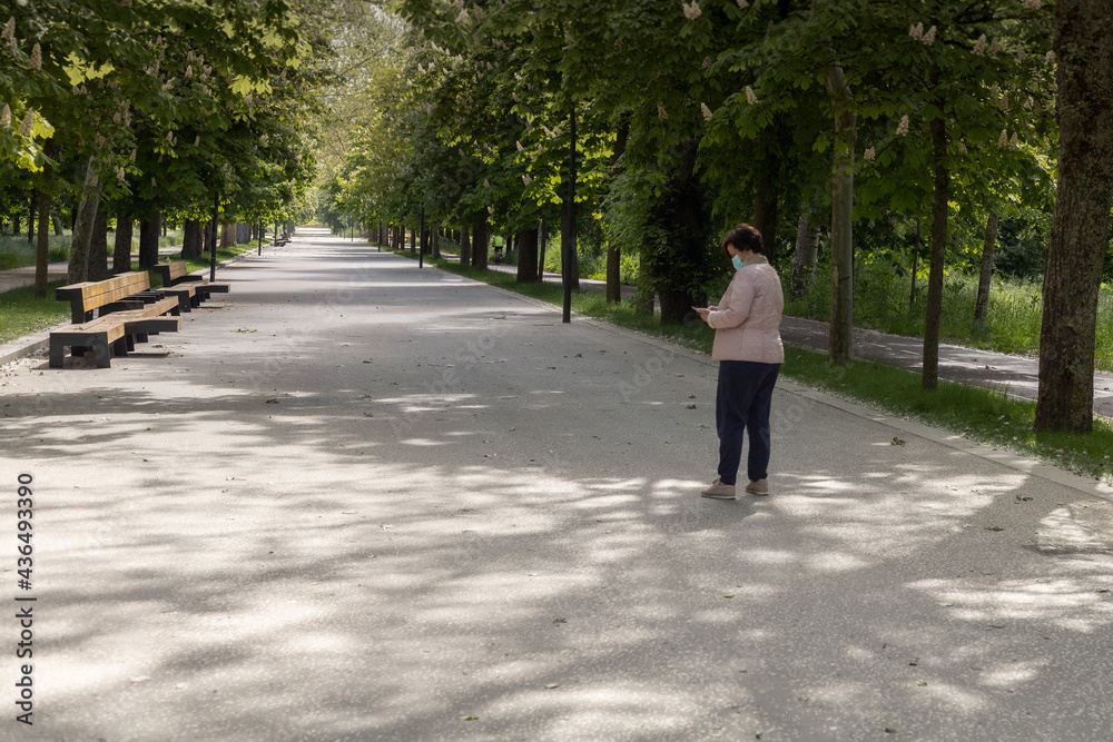 Middle-aged woman wearing a coronavirus mask texting on a smart phone while walking through a tree-lined park with a large path in a pink jacket