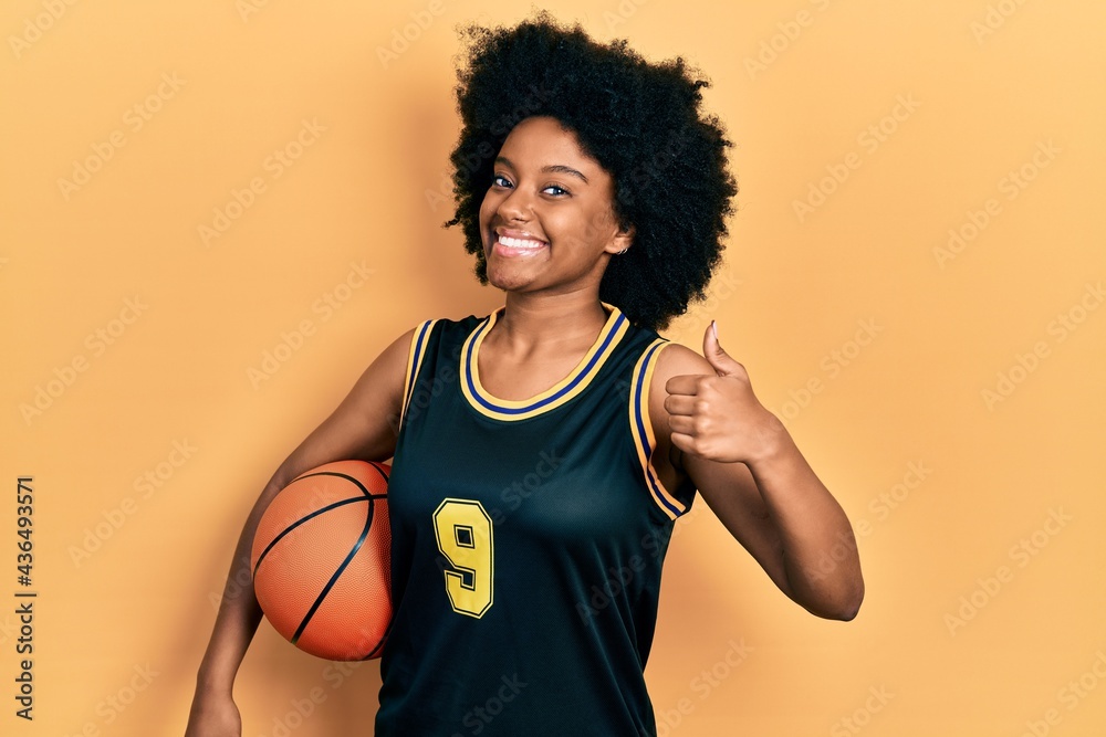 Young african american woman holding basketball ball smiling happy and positive, thumb up doing excellent and approval sign
