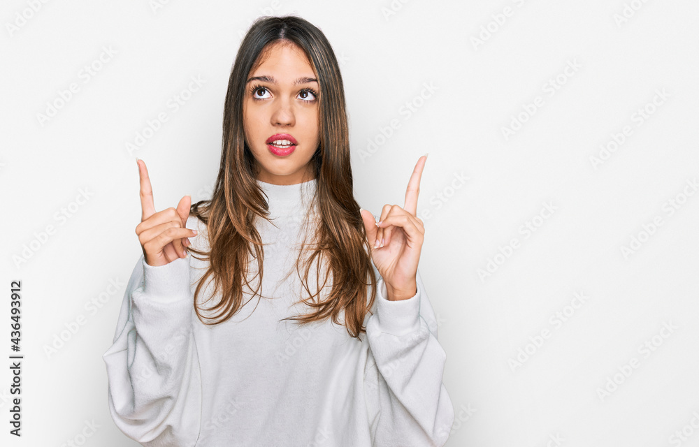 Young brunette woman wearing casual turtleneck sweater amazed and surprised looking up and pointing with fingers and raised arms. Young brunette woman wearing casual turtleneck sweater amazed and surprised looking up and pointing with fingers and raised arms.