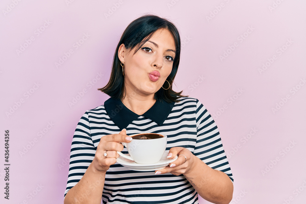 Young hispanic woman drinking a cup of coffee looking at the camera blowing a kiss being lovely and sexy. love expression. Young hispanic woman drinking a cup of coffee looking at the camera blowing a kiss being lovely and sexy. love expression.