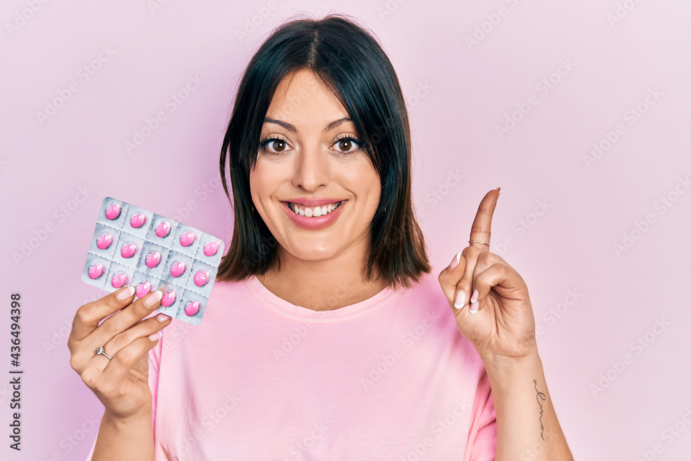 Young hispanic woman holding pills surprised with an idea or question pointing finger with happy face, number one Young hispanic woman holding pills surprised with an idea or question pointing finger with happy face, number one