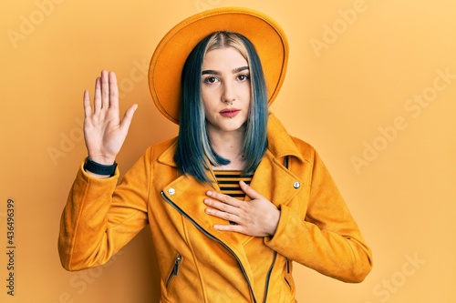 Young modern girl wearing yellow hat and leather jacket swearing with hand on chest and open palm, making a loyalty promise oath