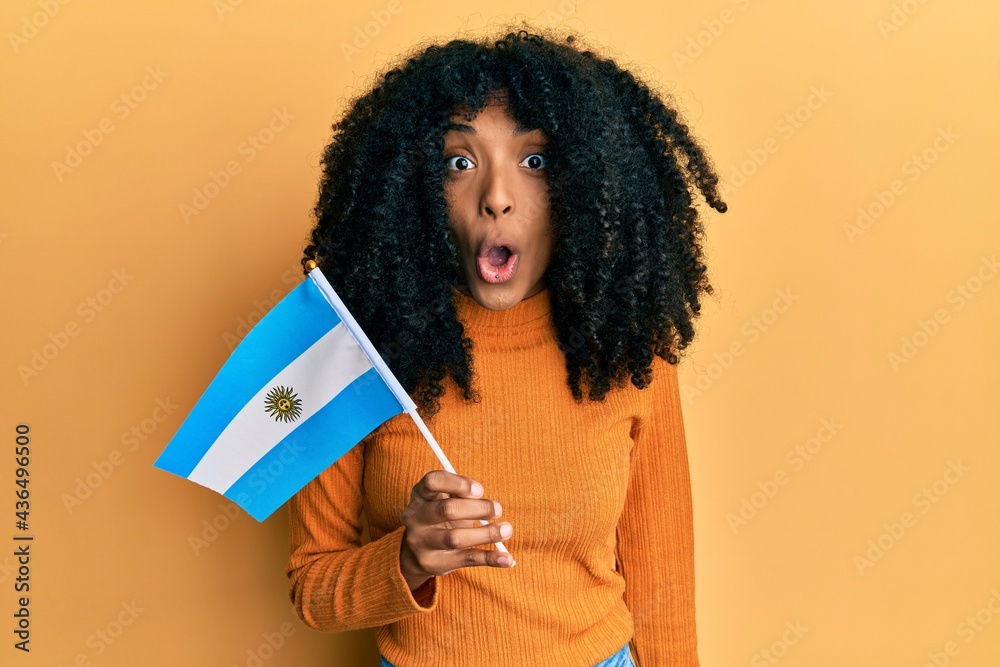 African american woman with afro hair holding argentina flag scared and amazed with open mouth for surprise, disbelief face African american woman with afro hair holding argentina flag scared and amazed with open mouth for surprise, disbelief face