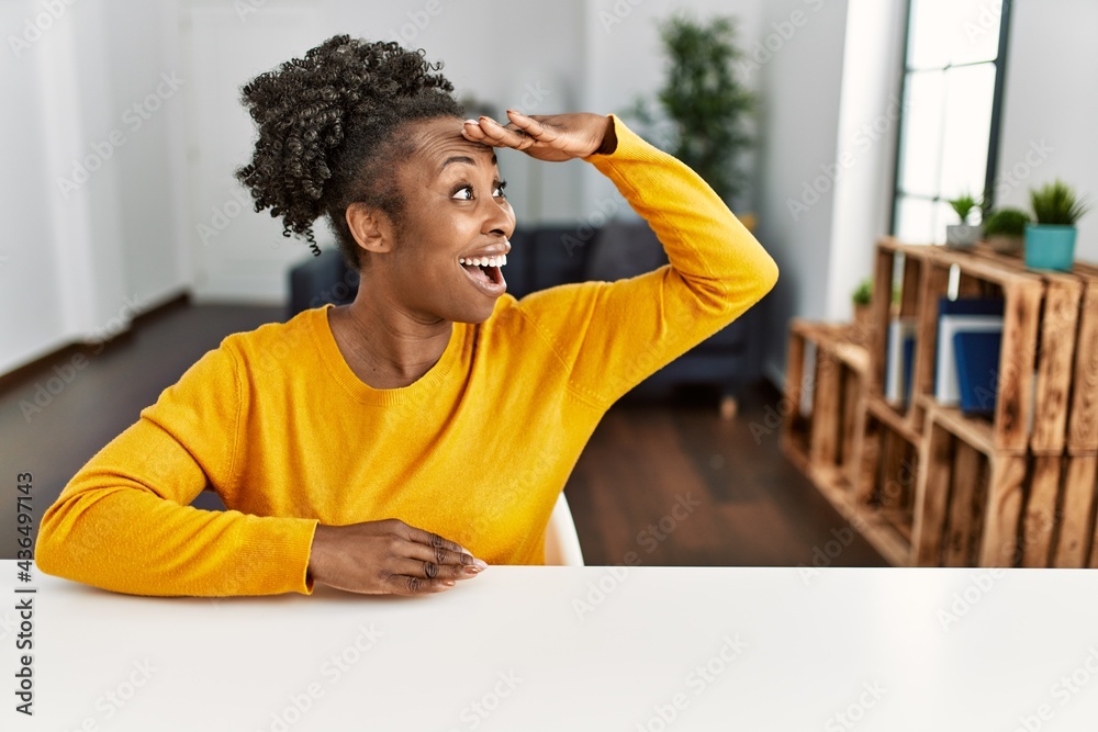 Young african american woman wearing casual clothes sitting on the table at home very happy and smiling looking far away with hand over head. searching concept.