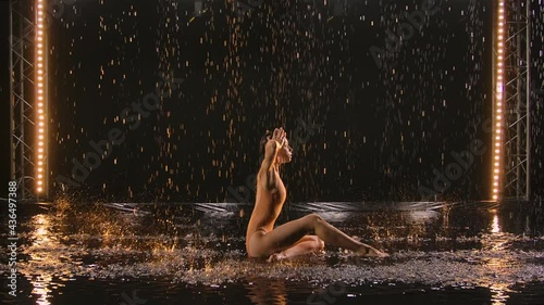 Young woman performs choreography modern ballet with acrobatic elements, bending body on water surface in rain. Silhouette of sexy female in wet swimsuit moves sensually in dark studio. Slow motion. photo