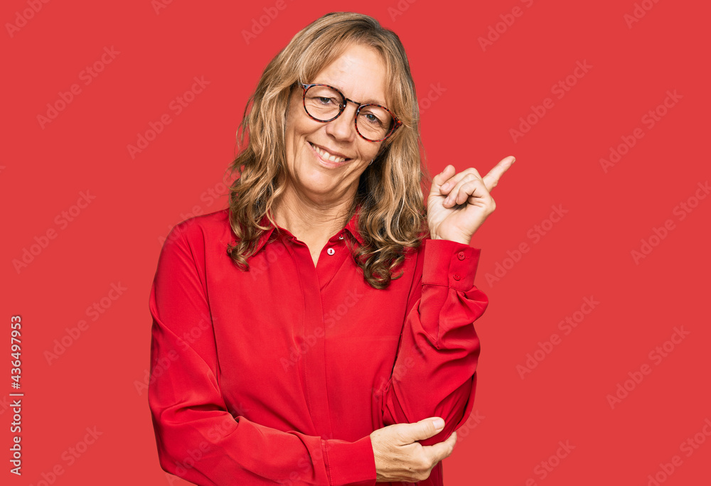 Middle age blonde woman wearing casual shirt over red background with a big smile on face, pointing with hand and finger to the side looking at the camera. Middle age blonde woman wearing casual shirt over red background with a big smile on face, pointing with hand and finger to the side looking at the camera.