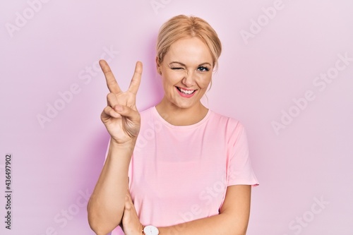 Young blonde woman wearing casual pink t shirt smiling with happy face winking at the camera doing victory sign with fingers. number two. © Krakenimages.com