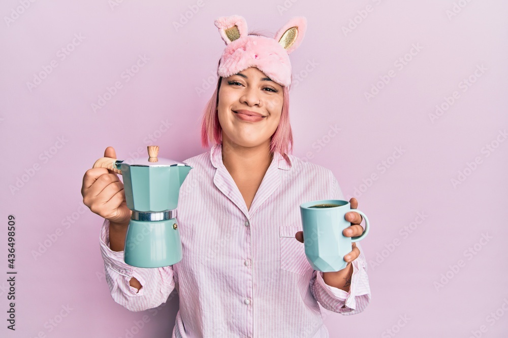 Hispanic woman with pink hair wearing sleep mask and pajama drinking a coffee cup smiling with a happy and cool smile on face. showing teeth. Hispanic woman with pink hair wearing sleep mask and pajama drinking a coffee cup smiling with a happy and cool smile on face. showing teeth.