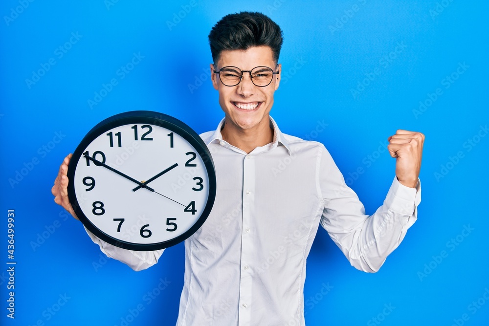 Young hispanic man holding big clock screaming proud, celebrating victory and success very excited with raised arm Young hispanic man holding big clock screaming proud, celebrating victory and success very excited with raised arm