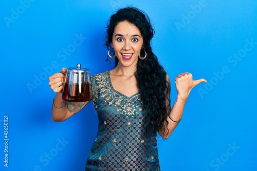 Young woman wearing bindi and traditional kurta holding traditional tea pot pointing thumb up to the side smiling happy with open mouth