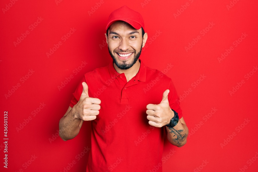 Hispanic man with beard wearing delivery uniform and cap success sign doing positive gesture with hand, thumbs up smiling and happy. cheerful expression and winner gesture.