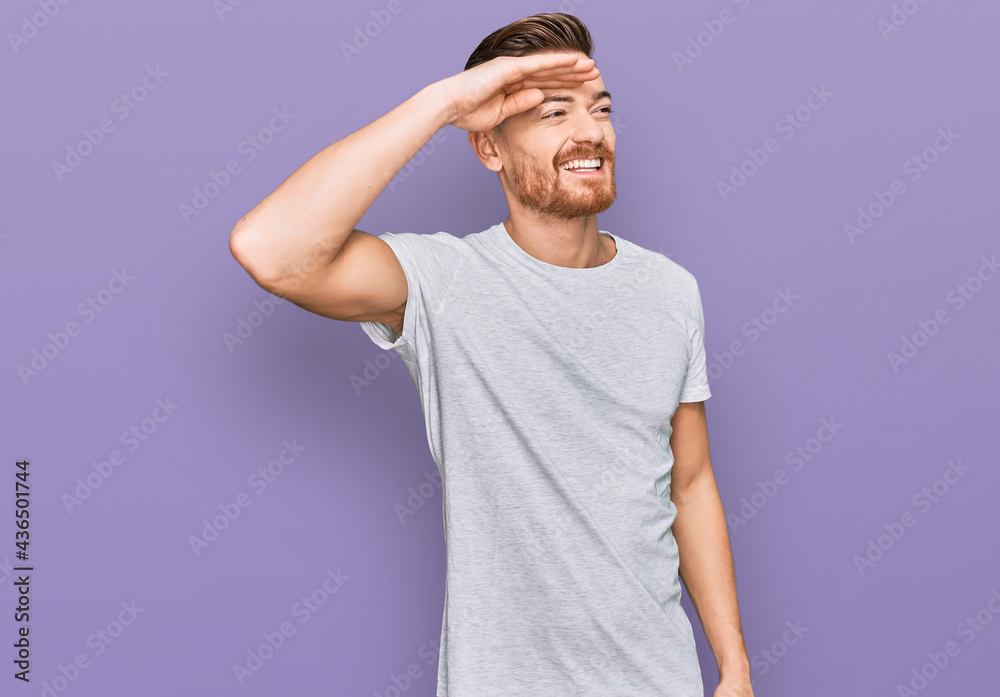 Young redhead man wearing casual grey t shirt very happy and smiling looking far away with hand over head. searching concept. Young redhead man wearing casual grey t shirt very happy and smiling looking far away with hand over head. searching concept.