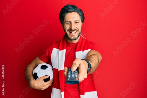 Cuadro en lienzo Young hispanic man football hooligan holding ball using tv control smiling and laughing hard out loud because funny crazy joke