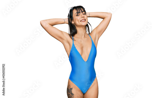 Young hispanic woman wearing swimsuit and sunglasses relaxing and stretching, arms and hands behind head and neck smiling happy