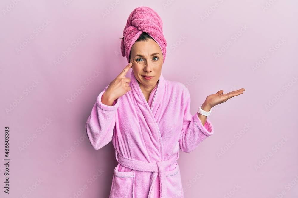 Young blonde woman wearing shower towel cap and bathrobe confused and annoyed with open palm showing copy space and pointing finger to forehead. think about it.